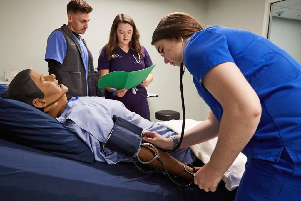 A P A student practices checking blood pressure on a patient simulator while two other students discuss the patient's health history
