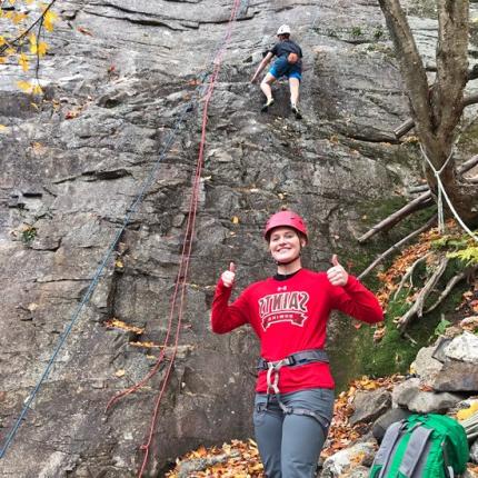A student gives two thumbs up in front of a mountainside that another student is rock climbing